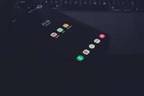 A smartphone lying face-up on a laptop keyboard with apps visible on its screen, including YouTube, Notes, and Gmail. The environment is dimly lit, creating a moody and tech-focused atmosphere.