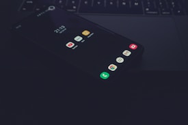 A smartphone lying face-up on a laptop keyboard with apps visible on its screen, including YouTube, Notes, and Gmail. The environment is dimly lit, creating a moody and tech-focused atmosphere.