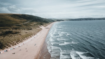 An expansive sandy beach stretches along the coastline, bordered by a lush green hillside. Small waves gently roll onto the shore, where a line of colorful beach huts is visible. A few people can be seen walking along the beach, enjoying the serene and open landscape under a partly cloudy sky.