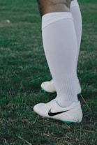 A close-up of a person wearing white knee-high socks and white sports shoes with a Nike logo. The individual stands on a grassy field.