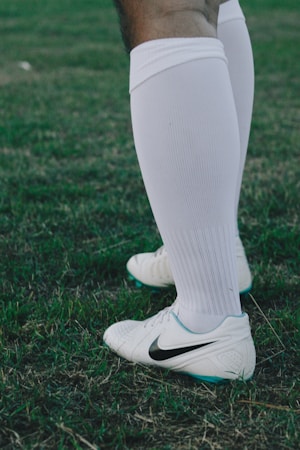 A close-up of a person wearing white knee-high socks and white sports shoes with a Nike logo. The individual stands on a grassy field.