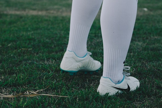 A pair of legs wearing white knee-high socks and white soccer cleats stand on a grassy field. The grass appears lush with a few dried blades scattered around.