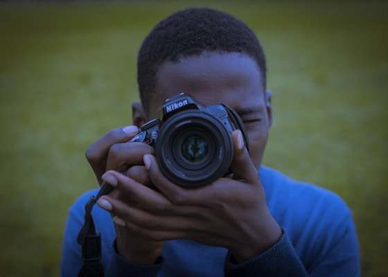 A person is holding a Nikon camera up to their eye, capturing a photograph. They are wearing a blue garment, and the background consists of out-of-focus greenery, suggesting an outdoor setting.