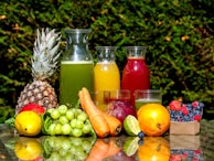 A selection of fresh fruits and vegetables is arranged on a reflective surface, including a pineapple, grapes, carrots, lime, orange, berries, and a red apple. There are also three glass carafes filled with colorful juices, possibly green, orange, and red, placed behind the fruits. The background is a lush green foliage.