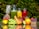 A selection of fresh fruits and vegetables is arranged on a reflective surface, including a pineapple, grapes, carrots, lime, orange, berries, and a red apple. There are also three glass carafes filled with colorful juices, possibly green, orange, and red, placed behind the fruits. The background is a lush green foliage.