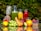 A selection of fresh fruits and vegetables is arranged on a reflective surface, including a pineapple, grapes, carrots, lime, orange, berries, and a red apple. There are also three glass carafes filled with colorful juices, possibly green, orange, and red, placed behind the fruits. The background is a lush green foliage.