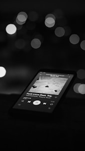 A smartphone displays a music player app with a song currently playing. The screen shows album art along with song details, and the background is blurred with bokeh lights, creating a soft and dimly lit atmosphere.