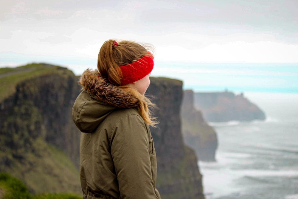 woman in brown jacket and red knit cap standing on cliff during daytime