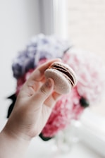 A close-up of a hand holding a macaron with a blurred background of a cozy café.