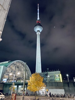 A tall television tower rises against a dark, cloudy night sky, illuminated with bright white lights and red accents. In the foreground, there is a bright yellow tree, a streetlamp, parked bicycles, and a modern building with a large curved glass roof.