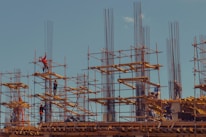 Workers are standing on steel scaffolding at a construction site, engaged in various tasks. The structure consists of wooden planks and metal rods reaching upward against a clear blue sky.
