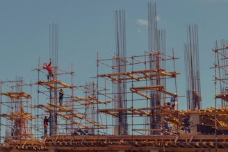 Workers are standing on steel scaffolding at a construction site, engaged in various tasks. The structure consists of wooden planks and metal rods reaching upward against a clear blue sky.