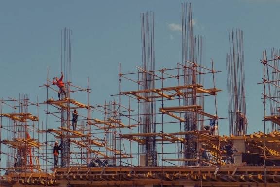 Workers are standing on steel scaffolding at a construction site, engaged in various tasks. The structure consists of wooden planks and metal rods reaching upward against a clear blue sky.