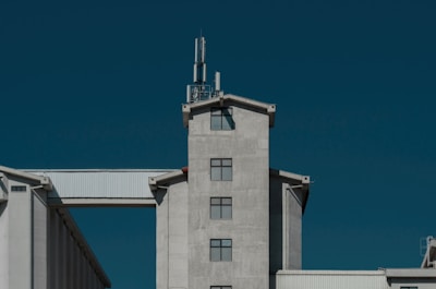 A large industrial building with a modern design and grey concrete facade is set against a clear, deep blue sky. It features a central tower with multiple square windows and a cellular antenna on top. A white, corrugated metal structure connects the tower to another part of the building.