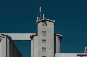 A large industrial building with a modern design and grey concrete facade is set against a clear, deep blue sky. It features a central tower with multiple square windows and a cellular antenna on top. A white, corrugated metal structure connects the tower to another part of the building.