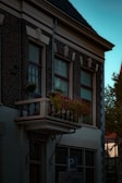 A classic European-style building with a small balcony adorned with a vibrant array of red flowers. The brickwork and ornate window design add to the historic charm. The building is situated under a serene blue sky.
