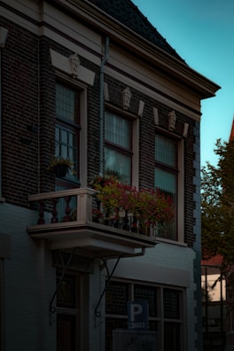 A classic European-style building with a small balcony adorned with a vibrant array of red flowers. The brickwork and ornate window design add to the historic charm. The building is situated under a serene blue sky.