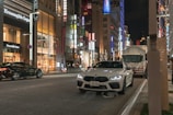 A bustling city street at night is lined with high-end stores, including Ermenegildo Zegna and Miu Miu. Bright store signs and decorative lights illuminate the scene. A white sports car is parked on the side of the road, while another car and a truck are visible on the street.