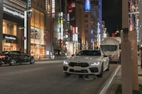 A bustling city street at night is lined with high-end stores, including Ermenegildo Zegna and Miu Miu. Bright store signs and decorative lights illuminate the scene. A white sports car is parked on the side of the road, while another car and a truck are visible on the street.