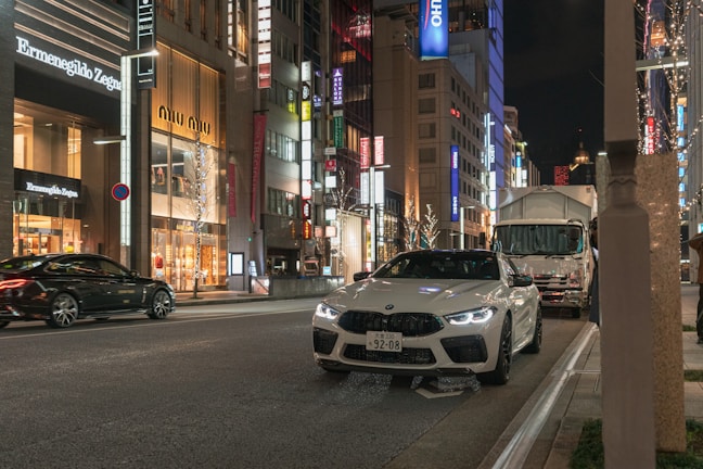 A bustling city street at night is lined with high-end stores, including Ermenegildo Zegna and Miu Miu. Bright store signs and decorative lights illuminate the scene. A white sports car is parked on the side of the road, while another car and a truck are visible on the street.