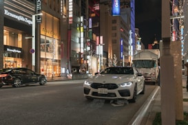 A bustling city street at night is lined with high-end stores, including Ermenegildo Zegna and Miu Miu. Bright store signs and decorative lights illuminate the scene. A white sports car is parked on the side of the road, while another car and a truck are visible on the street.