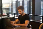 A smiling Indian man working remotely with a city skyline visible through the window behind him.
