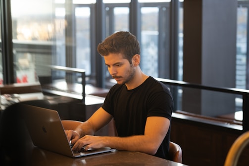 A man is working on a laptop in a modern indoor setting, with large windows offering a city view. He is seated at a table and focused on the screen.