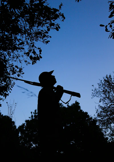 A rugged hunter silhouetted against a golden sunrise in a dense forest.