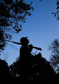 A silhouette of a person holding a rifle, surrounded by trees in a natural setting during twilight or dawn. The sky is a clear, deep blue with visible treetops and branches framing the scene.