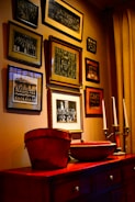 A wall adorned with multiple framed vintage photographs arranged in a gallery style, surrounded by a warm, ambient light. Below the frames is a wooden cabinet topped with decorative items including a red bucket and candlesticks.