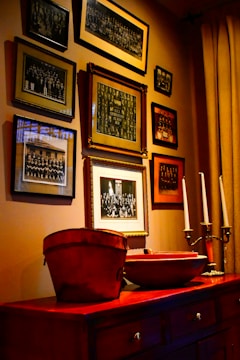 A wall adorned with multiple framed vintage photographs arranged in a gallery style, surrounded by a warm, ambient light. Below the frames is a wooden cabinet topped with decorative items including a red bucket and candlesticks.