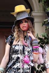 A person dressed in eclectic fashion, featuring multiple hats stacked on their head, vibrant and dramatic face makeup, and a sequin-covered outfit adorned with various accessories including a pink light bulb and strings of beads. The attire includes numerous bracelets and colorful bangles on their wrist.