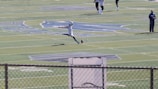 A football player in mid-action, kicking a ball on a large football field. The player is wearing sports gear, surrounded by a few other individuals scattered across the field. The field features distinct markings and logos, with bleachers visible in the background.
