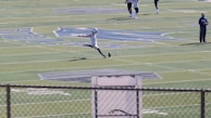 A football player in mid-action, kicking a ball on a large football field. The player is wearing sports gear, surrounded by a few other individuals scattered across the field. The field features distinct markings and logos, with bleachers visible in the background.