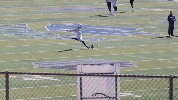 A football player in mid-action, kicking a ball on a large football field. The player is wearing sports gear, surrounded by a few other individuals scattered across the field. The field features distinct markings and logos, with bleachers visible in the background.