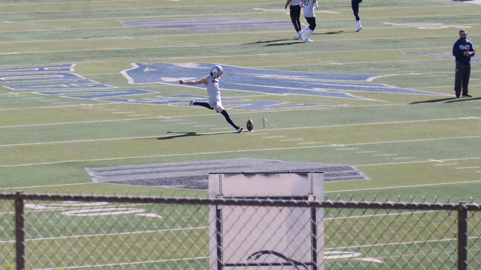 A football player in mid-action, kicking a ball on a large football field. The player is wearing sports gear, surrounded by a few other individuals scattered across the field. The field features distinct markings and logos, with bleachers visible in the background.