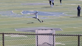 A football player in mid-action, kicking a ball on a large football field. The player is wearing sports gear, surrounded by a few other individuals scattered across the field. The field features distinct markings and logos, with bleachers visible in the background.