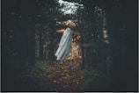 A ghostly figure draped in a white sheet hovers along a narrow forest path surrounded by dense, dark trees. The atmosphere is eerie, with minimal light filtering through the foliage.