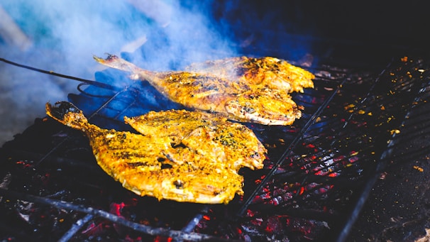 Three whole fish are being grilled on a barbecue. The fish have a golden-yellow marinade, likely containing spices and herbs, creating an appetizing appearance. Smoke rises from the grill, indicating they are being cooked over charcoal.