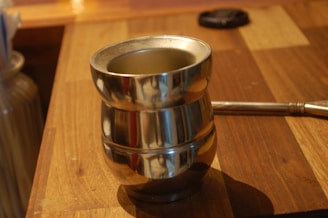 A metallic cylindrical container with a polished finish is placed on a wooden surface. The container has a reflective surface and a sturdy appearance with a narrow neck. In the background, part of a metal rod or utensil is visible.
