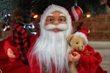 A festive Santa Claus figurine with a detailed face and white beard is dressed in a traditional red suit. It holds a small, brown teddy bear with a red ribbon, and a red and black checkered sack. The background features a decorated Christmas tree with ornaments and soft lighting.