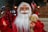 A festive Santa Claus figurine with a detailed face and white beard is dressed in a traditional red suit. It holds a small, brown teddy bear with a red ribbon, and a red and black checkered sack. The background features a decorated Christmas tree with ornaments and soft lighting.