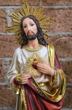 A religious statue depicting a figure with a serene expression, wearing a robe adorned with gold and red accents. The figure bears a sacred heart symbol on the chest, surrounded by a golden halo with sharp rays extending outward, set against a brick wall background.