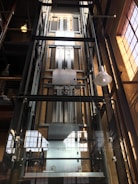 A modern glass elevator shaft inside a building with metal mechanisms and railings. The structure is enclosed with transparent panels, revealing the mechanical details. Sunlight streams in from large, industrial-style windows, creating a pattern of light and shadow.