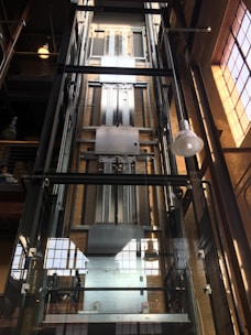 A modern glass elevator shaft inside a building with metal mechanisms and railings. The structure is enclosed with transparent panels, revealing the mechanical details. Sunlight streams in from large, industrial-style windows, creating a pattern of light and shadow.