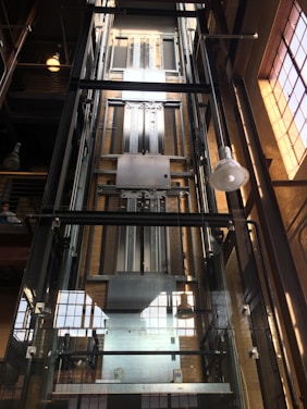 A modern glass elevator shaft inside a building with metal mechanisms and railings. The structure is enclosed with transparent panels, revealing the mechanical details. Sunlight streams in from large, industrial-style windows, creating a pattern of light and shadow.