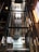A modern glass elevator shaft inside a building with metal mechanisms and railings. The structure is enclosed with transparent panels, revealing the mechanical details. Sunlight streams in from large, industrial-style windows, creating a pattern of light and shadow.