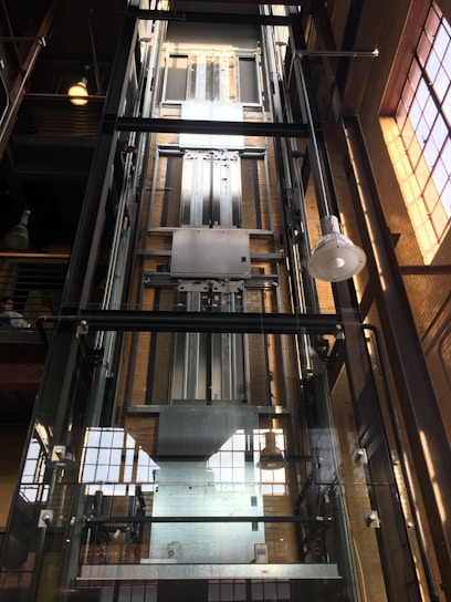 A modern glass elevator shaft inside a building with metal mechanisms and railings. The structure is enclosed with transparent panels, revealing the mechanical details. Sunlight streams in from large, industrial-style windows, creating a pattern of light and shadow.