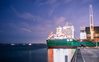 A large cargo ship is docked at a harbor during dusk, with bright lights illuminating the deck. The background includes a softly lit sky with stars and a visible streak of light, possibly from an aircraft or shooting star. A walkway extends along the harbor, and there are blurred figures and an umbrella on the side, suggesting movement or long exposure photography.