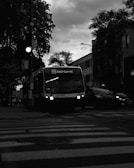 A black and white image of a bus on route 55 Saint-Laurent in a city setting. The bus is positioned near a pedestrian crossing and surrounded by buildings and trees. Streetlights and car headlights are visible, with a gloomy, overcast sky in the background.
